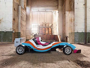 Ein Rennwagen mit auffälligem Design in Blau, Orange und Pink steht in einer verlassenen Lagerhalle. Die Wände sind aus Beton, und Licht dringt durch Fenster im Hintergrund. Die Atmosphäre ist industriell und leer. Der Wagen hat die Nummer 62.