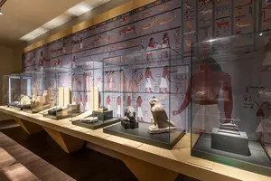 Die Abbildung zeigt eine Museumsansicht mit Vitrinen, die verschiedene antike Skulpturen und Ausstellungsstücke präsentieren. Im Hintergrund ist eine Wandmalerei mit ägyptischen Motiven zu sehen. Die Vitrinen sind sorgfältig illuminiert und präsentieren artefakte aus der alten ägyptischen Kultur.