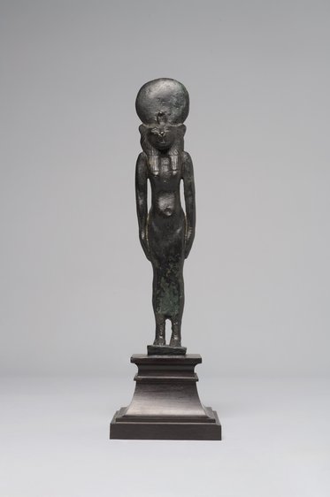 Eine bronze Statue einer stehenden Frau, die ein hohes Kopfbedeckung trägt. Sie hat eine schlanke Figur und ist auf einem rechteckigen Sockel platziert. Die Oberfläche der Statue zeigt Patina und Alterserscheinungen. Der Hintergrund ist einfarbig hell.
