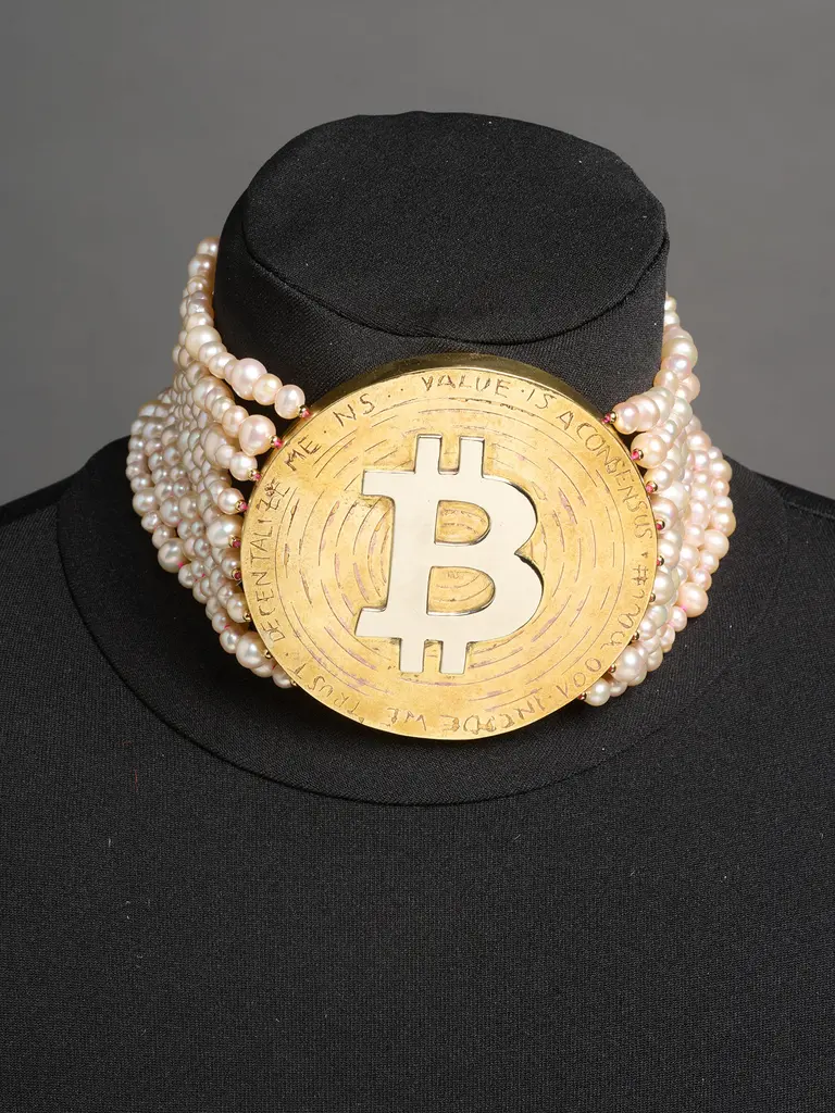 A creative piece of jewelry featuring a large, gold-colored Bitcoin symbol as a pendant, surrounded by strands of white pearls. The necklace is displayed on a black mannequin bust, emphasizing its unique blend of modern cryptocurrency and classic elegance.