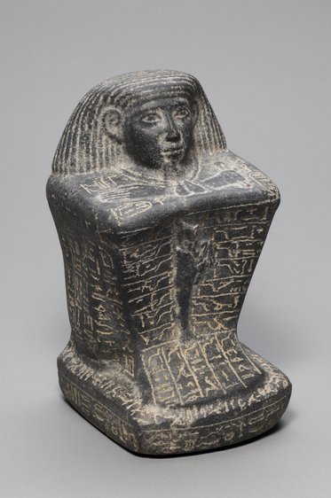 Eine schwarze ägyptische Statuette mit einem menschlichen Kopf und geflochtenen Haaren, auf einem rechteckigen Sockel. Die Oberfläche ist mit Hieroglyphen verziert, die bis zur unteren Kante reichen. Die Figur steht auf einem neutralen Hintergrund.