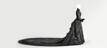 Eine schwarze, elegante Robe mit langem, geschwungenem Saum, die eine silberne mannequinähnliche Figurenstellung zeigt. Die Detailverarbeitung der Robe umfasst feine Muster und Texturen, die auf die Kleiderkunst hinweisen. Der Hintergrund ist neutral und hebt die Robe hervor.