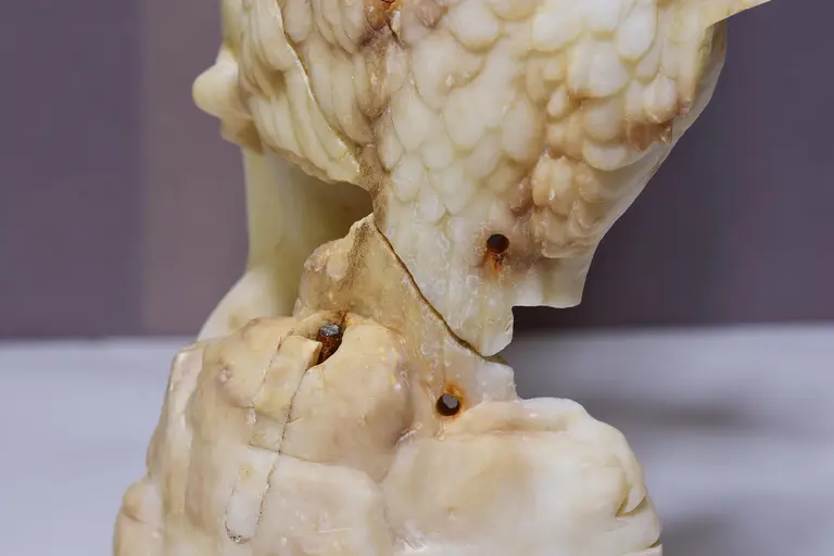 Eine kunstvolle Skulptur aus Onyx, die einen Vogel darstellt. Der Vogel sitzt auf einem geschwungenen Sockel, sichtbar sind feine Details im Gefieder und die Struktur des Steins. Die Farbnuancen variieren von Beige bis Bernstein.