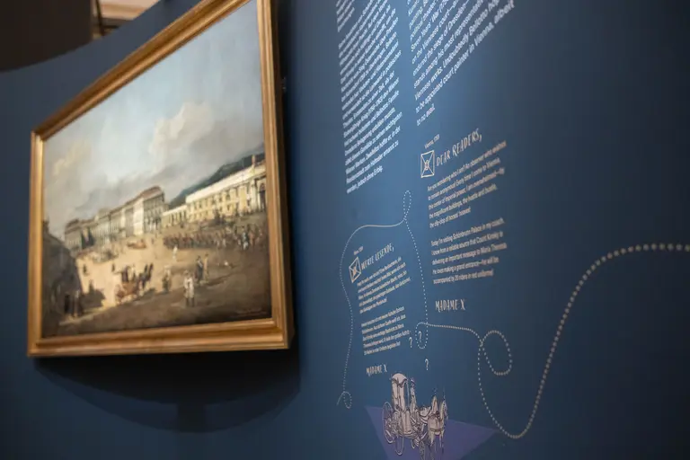 Ein Gemälde in einem goldenen Rahmen zeigt eine belebte Stadtansicht mit Menschen und Gebäuden. Im Hintergrund befindet sich eine dunkle Wand mit informativem Text und Illustrationen, die das Bild und seine Bedeutung erklären.