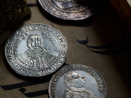 Das Bild zeigt mehrere historische silberne Münzen auf einem alten Dokument. Eine Münze ist besonders gut sichtbar und zeigt das Portrait eines Herrschers mit reich verzierten Kleidern und einem Kragen. Die Münzen sind auf einem dunklen Hintergrund platziert.