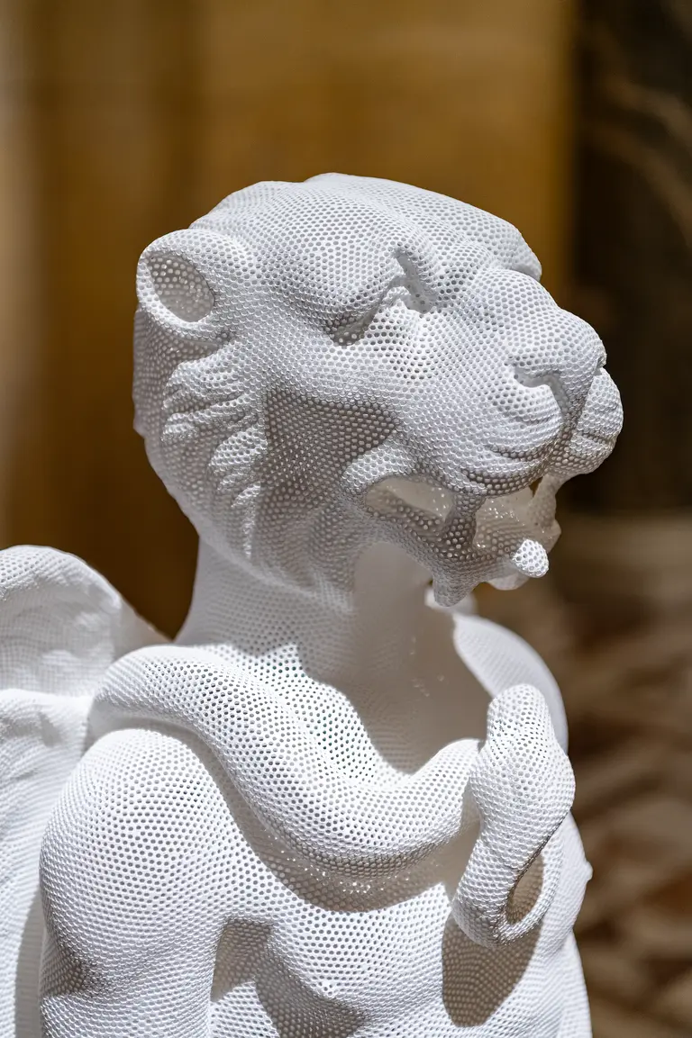 Skulptur eines Löwen mit schlängelndem Körper, aus einem feinen, weißen Netzmaterial gefertigt. Die Statue zeigt detaillierte Gesichtszüge und ist im Vordergrund platziert, während der Hintergrund unscharf bleibt.