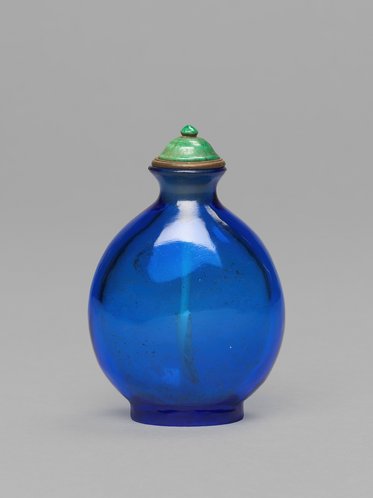 Eine runde, blaue Flasche aus Glas mit einem grünen Deckel. Die Flasche hat eine glatte Oberfläche und eine leicht geschwungene Form. Der Deckel hat eine kleine, glänzende grüne Verzierungen.