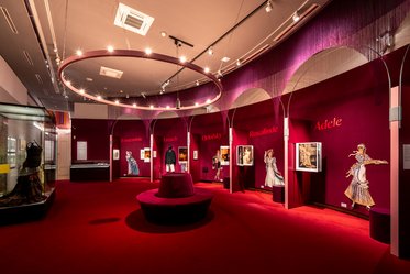 Eine Ausstellungsansicht mit rot gefärbten Wänden, auf denen verschiedene Namen stehen. In der Mitte steht eine runde Plattform mit einer Sammlung von Kostümen und Bildern aus einer Theater- oder Modeausstellung, die das Thema Vielfalt und Geschichte des Theaters betont.
