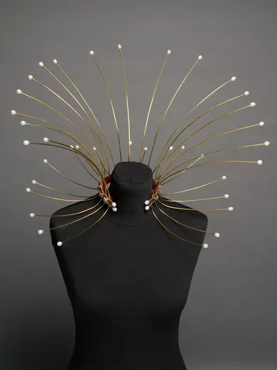 A unique, artistic collar displayed on a black mannequin. The collar features thin, golden rods radiating outward, adorned with delicate pearls at the tips, creating a striking, modern look. The background is a simple gray, emphasizing the collar's design.