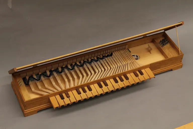 Fotografie eines historischen Clavichords: Das Instrument ist vollständig geöffnet, wodurch die innere Mechanik sichtbar wird und steht vor einem neutralen grauen Hintergrund. Die Holzkonstruktion zeigt eine schlichte, handwerklich präzise Verarbeitung. Vorne befinden sich die Tasten aus hellem Holz, mit dunklen, erhöhten Halbtontasten. Über den Tasten sind die Tangenten sichtbar, die beim Drücken der Tasten die Saiten anschlagen. Die Saiten sind über einen Resonanzboden gespannt, der ein dekoratives, rundes Schallloch mit einem ornamentalen Muster aufweist. Rechts im Bild ist der Bereich für die Befestigung der Saiten zu sehen.