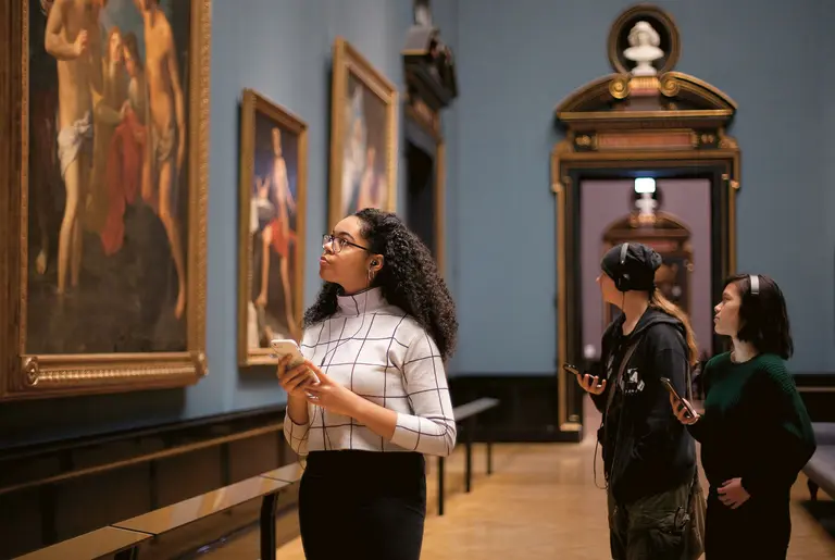 In einem Museum betrachten drei Personen Kunstwerke an den Wänden. Eine Frau mit lockigem Haar und Brille hält ein Smartphone und hört aufmerksam zu. Die anderen zwei Personen, eine in schwarzer Mütze und eine in grünem Pullover, halten ebenfalls Smartphones und scheinen interessiert.