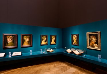 Ein Ausstellungsraum mit einer blauen Wand, die sechs Porträts und ein größeres Gemälde zeigt. In der Mitte stehen zwei Bücher auf einem Tisch. Die Kunstwerke stellen Personen mit Blumen und unterschiedlichen Ausdrücken dar. Der Boden ist aus Holz mit einem Fischgrätenmuster.
