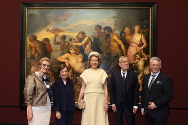 Fünf Personen posieren in eleganter Kleidung vor einem großen, historischen Gemälde, das mythologische Szenen mit nackten Figuren darstellt. Der Hintergrund ist rot und der Rahmen des Gemäldes ist dunkel gehalten. Die Gruppe steht mittig, lächelt und blickt zur Kamera.