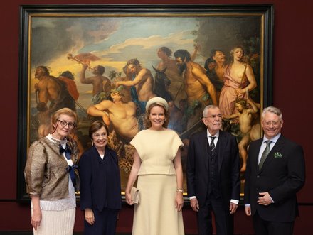 Fünf Personen posieren in eleganter Kleidung vor einem großen, historischen Gemälde, das mythologische Szenen mit nackten Figuren darstellt. Der Hintergrund ist rot und der Rahmen des Gemäldes ist dunkel gehalten. Die Gruppe steht mittig, lächelt und blickt zur Kamera.