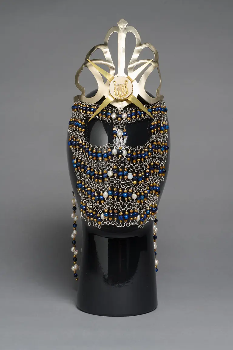 A decorative headpiece displayed on a mannequin head, featuring intricate beadwork in blue and gold, embellished with pearls. The design includes a prominent golden crown-like structure at the top. The headpiece combines elegance and artistry, showcasing craftsmanship in its detailed arrangement.