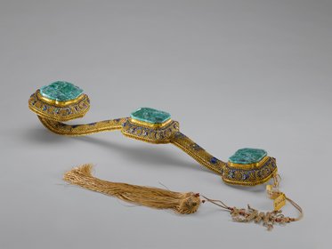 Ein kunstvoll gestalteter Ziergürtel mit drei rechteckigen, grünen Edelsteinen, umgeben von goldfarbenem und buntem Metall. Am Ende des Gürtels hängt ein längerer, geflochtener Faden mit kleinen Perlen. Der Gürtel zeigt extravagante Handwerkskunst und reich verzierte Details.