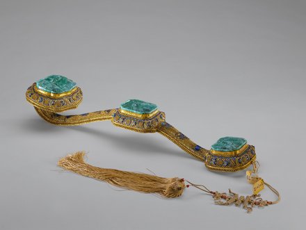 Ein kunstvoll gestalteter Ziergürtel mit drei rechteckigen, grünen Edelsteinen, umgeben von goldfarbenem und buntem Metall. Am Ende des Gürtels hängt ein längerer, geflochtener Faden mit kleinen Perlen. Der Gürtel zeigt extravagante Handwerkskunst und reich verzierte Details.