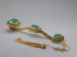 Ein kunstvoll gestalteter Ziergürtel mit drei rechteckigen, grünen Edelsteinen, umgeben von goldfarbenem und buntem Metall. Am Ende des Gürtels hängt ein längerer, geflochtener Faden mit kleinen Perlen. Der Gürtel zeigt extravagante Handwerkskunst und reich verzierte Details.