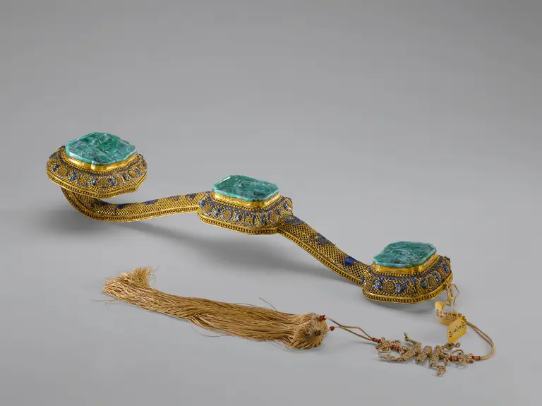 Ein kunstvoll gestalteter Ziergürtel mit drei rechteckigen, grünen Edelsteinen, umgeben von goldfarbenem und buntem Metall. Am Ende des Gürtels hängt ein längerer, geflochtener Faden mit kleinen Perlen. Der Gürtel zeigt extravagante Handwerkskunst und reich verzierte Details.