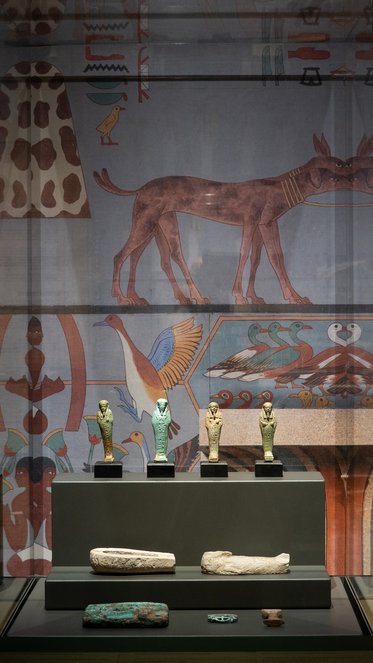 Alttext: Im Vordergrund stehen fünf kleine Statuen aus verschiedenen Materialien auf einem Podest. Im Hintergrund ist eine Wandmalerei mit Tieren und symbolischen Motiven aus dem alten Ägypten zu sehen.