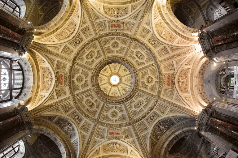 Ein Blick auf die kunstvoll gestaltete Decke eines Gebäudes, die mit goldenen Ornamenten und detaillierten Mustern verziert ist. Im Zentrum befindet sich ein kreisrunder Ausblick, durch den Licht strömt, und die Wände sind von Säulen flankiert.