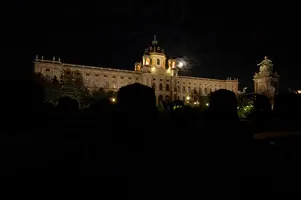 Das Bild zeigt ein eindrucksvolles historisches Gebäude in der Nacht, beleuchtet von warmem Licht. Der Mond ist teilweise sichtbar und trägt zur Atmosphäre bei. Um das Gebäude sind dunkelgrüne Hecken und ein schattiger Park zu sehen.