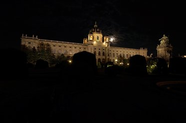 Das Bild zeigt ein eindrucksvolles historisches Gebäude in der Nacht, beleuchtet von warmem Licht. Der Mond ist teilweise sichtbar und trägt zur Atmosphäre bei. Um das Gebäude sind dunkelgrüne Hecken und ein schattiger Park zu sehen.