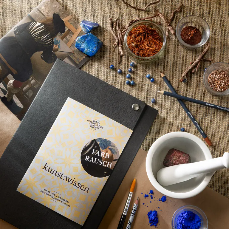 Eine Ansammlung von Kunstmaterialien auf einem Tisch. Im Vordergrund liegt ein schwarzes Heft mit dem Titel 