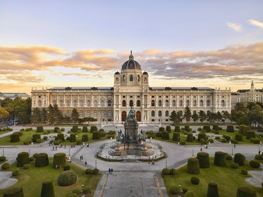 Das Bild zeigt das Kunsthistorische Museum in Wien, umgeben von gepflegten Gärten und Hecken. Im Vordergrund befindet sich ein großzügiger Brunnen, der von Menschen umgeben ist, während der Himmel in warmen Farben erleuchtet ist. Die Architektur des Museums ist beeindruckend und historisch.