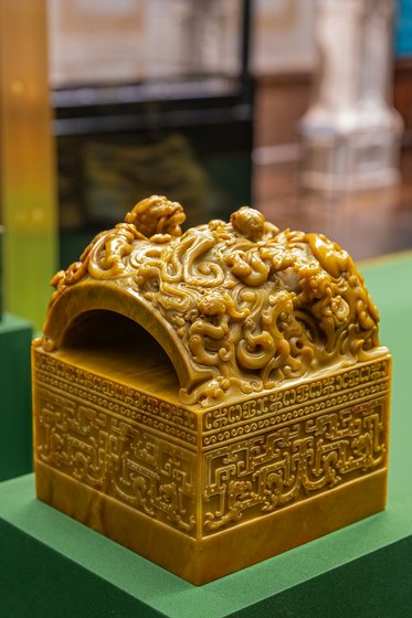 Ein kunstvoll geschnitzter, gelber Jadebehälter mit einem gewölbten Deckel, verziert mit detaillierten reliefartigen Figuren. Die Sockelstruktur zeigt feine Muster. Der Behälter steht auf einer grünen Oberfläche und hebt sich durch seine glatte, glänzende Oberfläche hervor.