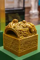 Ein kunstvoll geschnitzter, gelber Jadebehälter mit einem gewölbten Deckel, verziert mit detaillierten reliefartigen Figuren. Die Sockelstruktur zeigt feine Muster. Der Behälter steht auf einer grünen Oberfläche und hebt sich durch seine glatte, glänzende Oberfläche hervor.