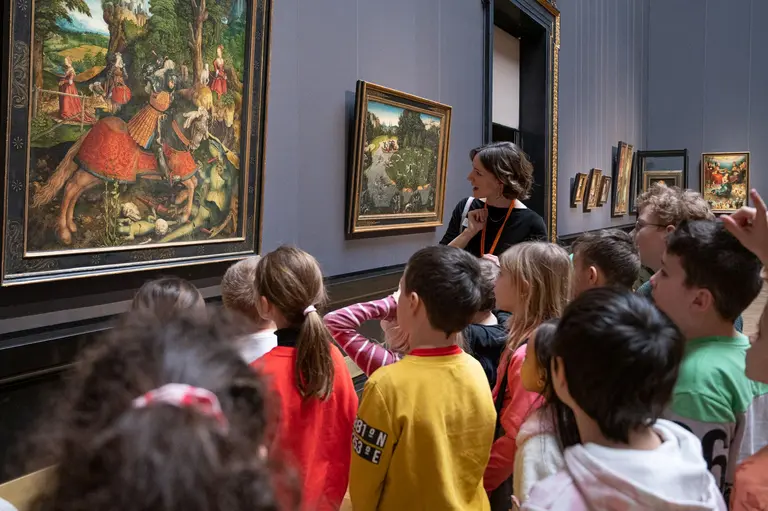 Eine Lehrerin erklärt einer Gruppe von Kindern ein Gemälde in einem Museum. Die Kinder schauen aufmerksam auf das Kunstwerk, während sie zuhören. Das Bild zeigt eine historische Szene mit mehreren Reitern.