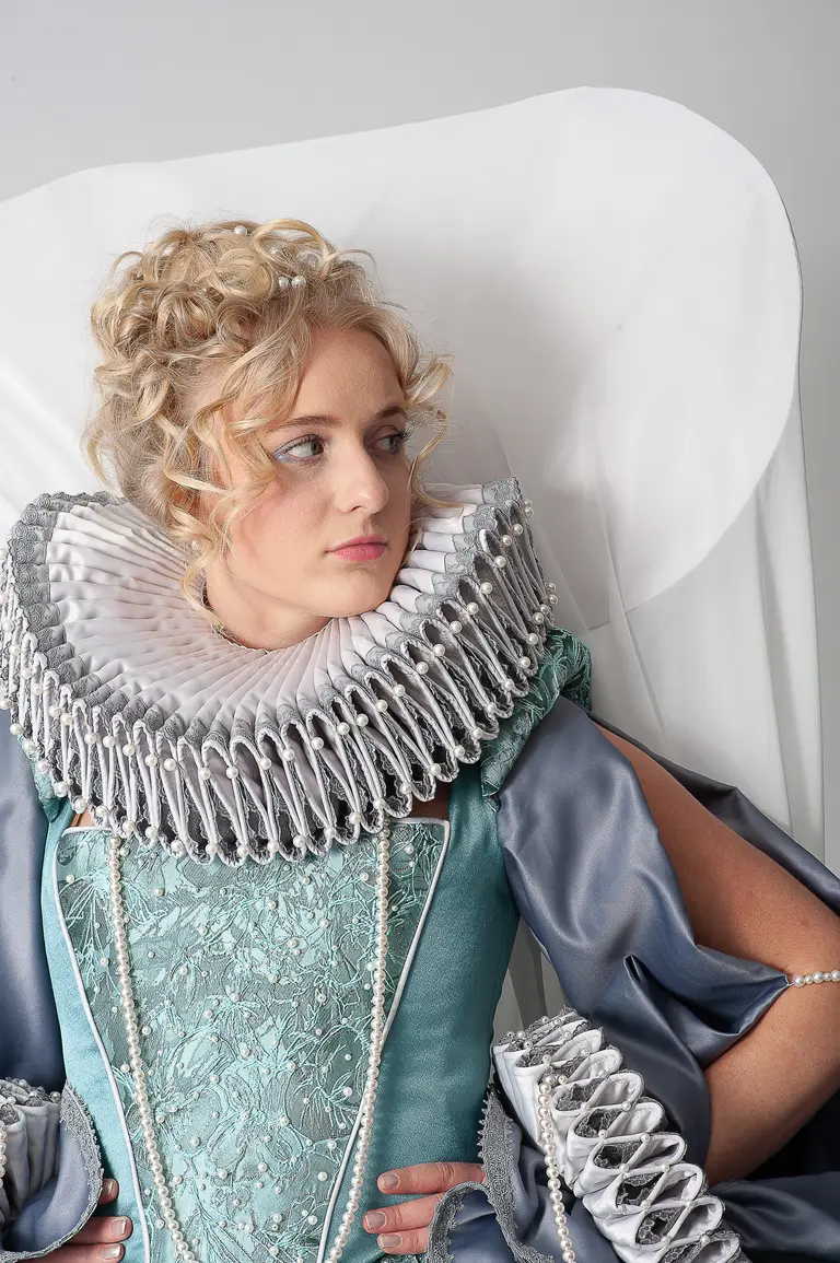 A young woman with curly hair wears an elaborate historical costume featuring a turquoise bodice and a large, ruffled collar. She poses thoughtfully, with her arms crossed and a serious expression, against a light background that adds to the dramatic effect of her attire.