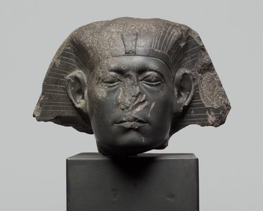 Eine antike, beschädigte Kopfskulptur eines Pharaos aus schwarzem Stein. Der Pharao hat eine charakteristische Frisur mit Flügeln und trägt ein Kopfband. Der Gesichtsausdruck ist neutral, jedoch sind einige Details, wie die Nase, stark abgetragen. Die Statue steht auf einem schlichten Sockel.
