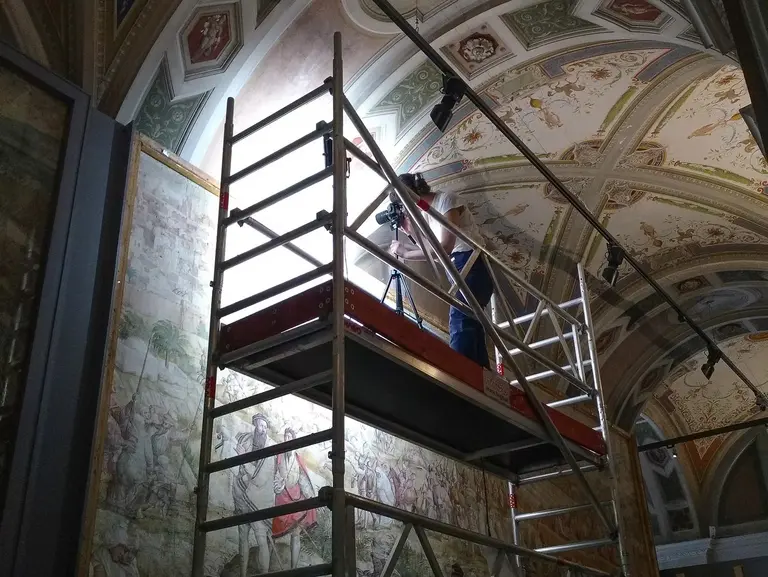 Ein Restaurator steht auf einem hohen Gerüst und fotografiert ein Fresko an der Decke. Das Gewölbe ist mit bunten Ornamenten und Malereien verziert. An der Wand unter dem Gerüst befindet sich der Vermeyen Karton.