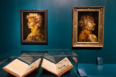 Zwei Gemälde an der Wand, eines zeigt eine figurative Darstellung mit einer aufwendigen Kopfdekoration, das andere einen Mensch mit einem Tierkopf. Vor den Gemälden befindet sich eine Vitrine mit offenen alten Büchern und Beschilderung. Der Hintergrund ist in einem tiefen Blau gehalten.