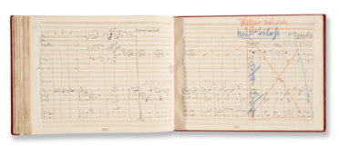 Das Bild zeigt eine offene Partitur in einem Notizbuch mit handschriftlichen Musiknoten. Auf der rechten Seite sind durchgestrichene Textpassagen und Anmerkungen mit blauer und roter Tinte zu sehen, die Anweisungen für die Aufführung geben.