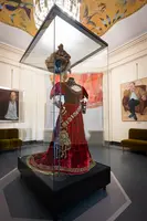 Ein prachtvolles, rotes Gewand mit aufwendigen Verzierungen steht in einem Glaskasten. Das Outfit wird von einer Krone ergänzt und ist Teil einer Ausstellung, umgeben von gemalten Portraits und einem eleganten Raumdesign.