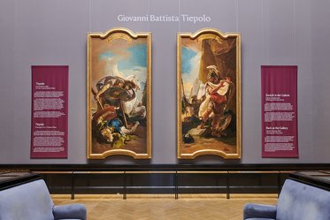 Zwei große Gemälde von Giovanni Battista Tiepolo hängen an einer Wand in einer Galerie. Links ist eine Szene mit einem Kämpfer und einem Schild, rechts ist ein männlicher Charakter in Rüstung dargestellt. An der Wand sind auch informative Tafeln über die Werke angebracht.