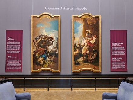 Zwei große Gemälde von Giovanni Battista Tiepolo hängen an einer Wand in einer Galerie. Links ist eine Szene mit einem Kämpfer und einem Schild, rechts ist ein männlicher Charakter in Rüstung dargestellt. An der Wand sind auch informative Tafeln über die Werke angebracht.