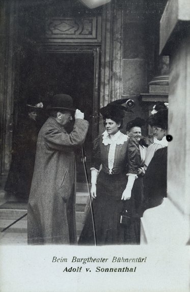 Eine monochrome Fotografie zeigt eine Gruppe von Menschen vor dem Burgtheater in Wien. Ein Mann mit Hut spricht mit einer elegant gekleideten Frau, während andere Frauen in der Hintergrund stehen. Die Atmosphäre wirkt feierlich und gesellschaftlich.
