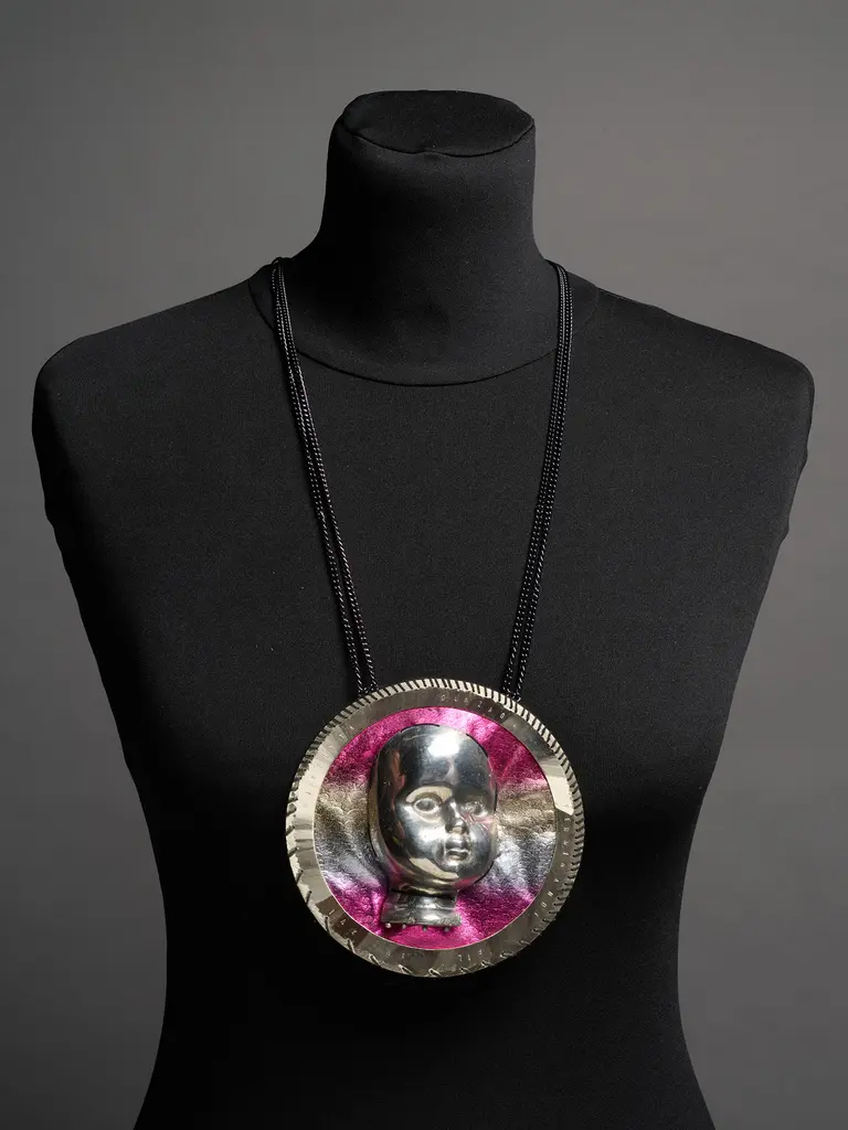 A mannequin displays a large, circular pendant necklace featuring a raised face design in metallic silver and colorful hues of pink. The necklace is attached with a simple black cord, highlighting the artistic craftsmanship of the pendant. The background is neutral to emphasize the jewelry.