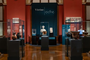 Ein Ausstellungsraum mit beleuchteten Vitrinen, die antike Kunstgegenstände wie Töpferwaren und Skulpturen präsentieren. Im Hintergrund steht ein erhobenes Display mit einer Statue und der Aufschrift „Vitrine extra“ an einer roten Wand.