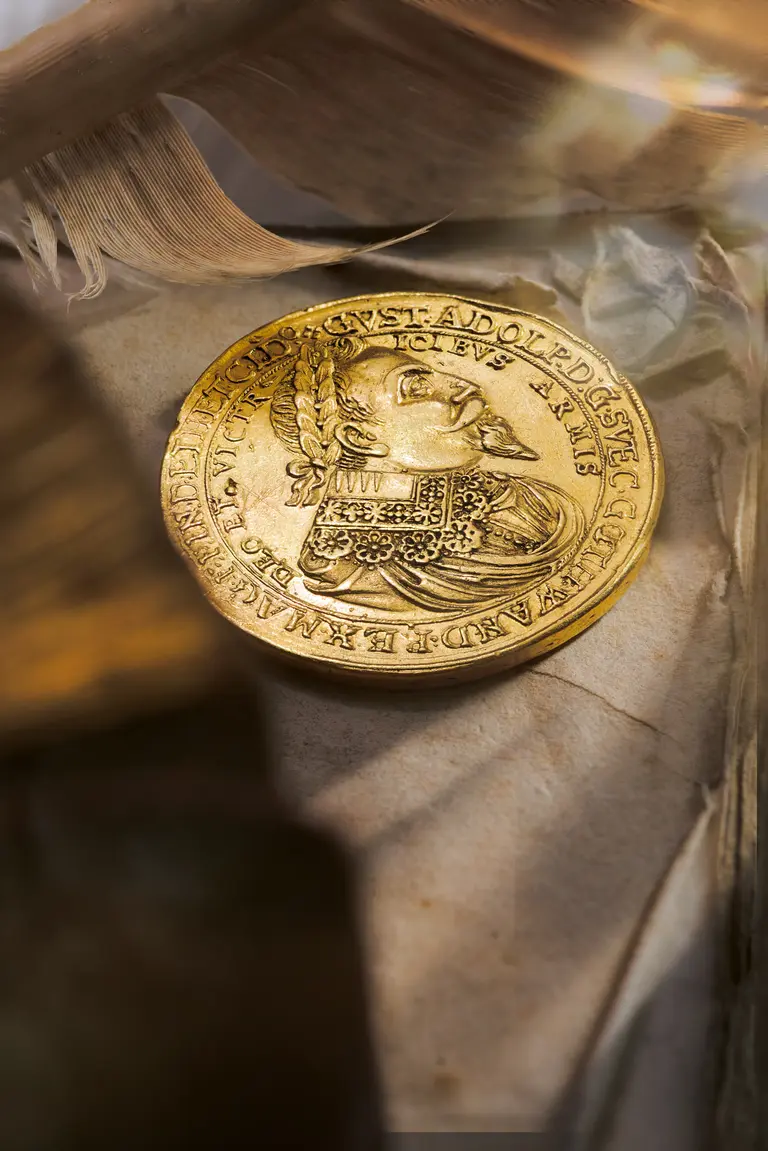 A gold coin is prominently displayed, featuring a detailed profile of a historical figure adorned with a collar and elaborate hair. The coin is partially covered by crumpled paper, hinting at its age and historical significance. Soft lighting highlights its shine and details.