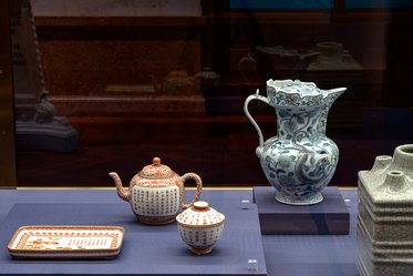 Auf dem Bild sind mehrere kunstvolle Keramiken zu sehen. Im Vordergrund stehen eine Teekanne, eine kleine Schüssel und ein Teller, die alle mit feinen Mustern verziert sind. Im Hintergrund befindet sich eine größere Kanne mit buntem, geschwungenem Design.