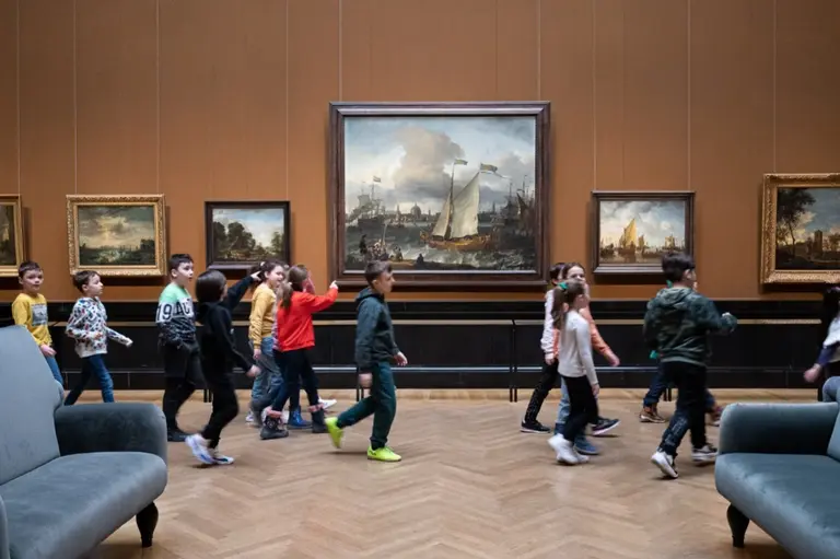 Eine Gruppe von Kindern bewegt sich durch ein Museum. Im Hintergrund sind mehrere Gemälde an der Wand zu sehen, darunter ein größeres Landschaftsgemälde mit Schiffen. Die Kinder tragen bunte Kleidung und scheinen interessiert an den Kunstwerken.