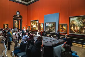 Ein Kunstführer spricht vor einer Gruppe von Menschen in einem Museum. Die Wände sind orange, und mehrere Gemälde hängen ausgestellt. Die Besucher sind aufmerksam und sitzen zum Teil auf einer Sitzgelegenheit im Vordergrund.