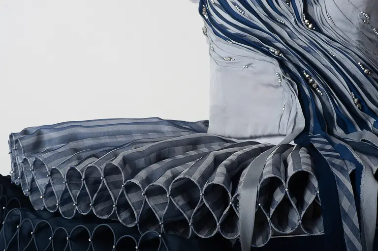 Close-up of a layered fabric garment featuring intricate pleats and ribbon details in shades of blue. The design includes decorative embellishments and a structured silhouette, showcasing the craftsmanship and texture of the fabric.