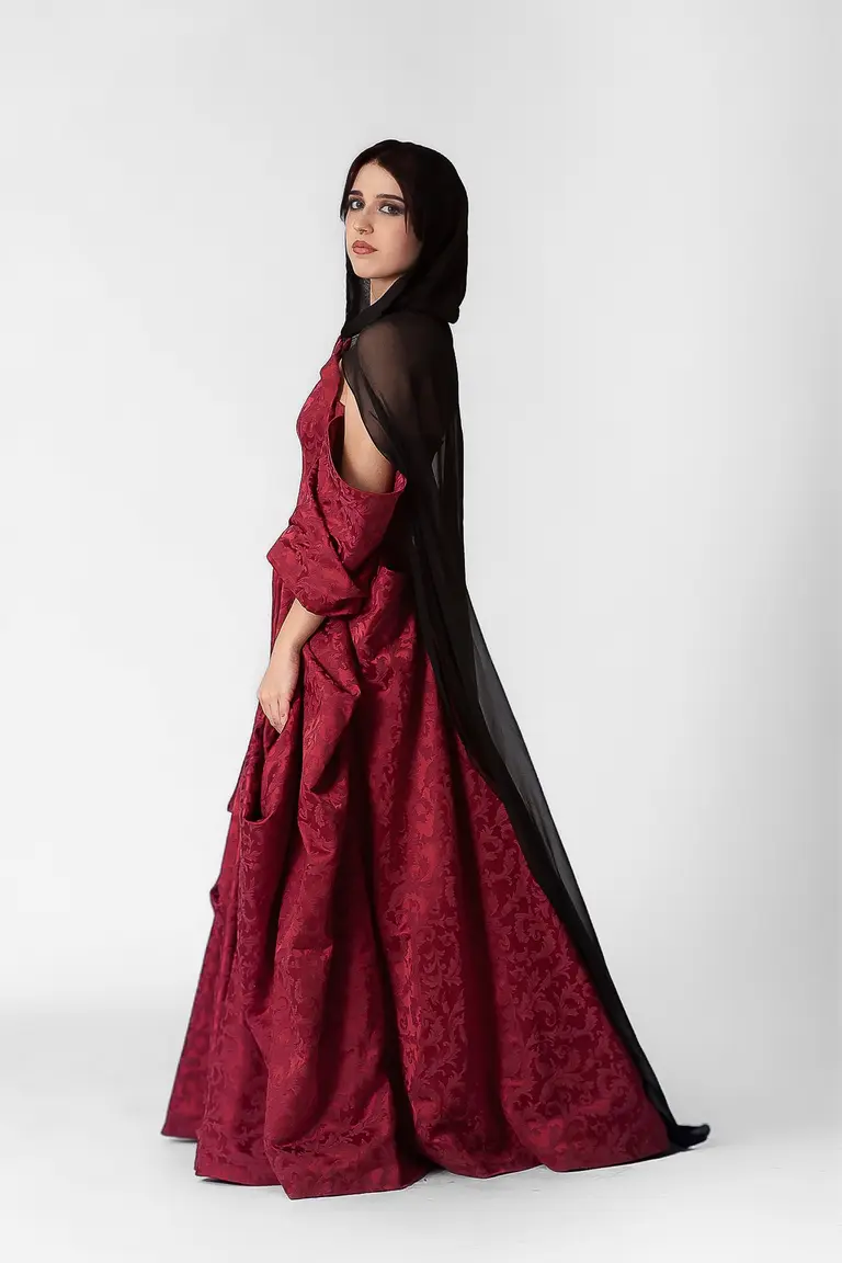 A young woman stands elegantly in profile, wearing a rich red evening gown with intricate patterns and a flowing black sheer cape. She gazes thoughtfully to the side, highlighting the gown's voluminous fabric and her graceful posture against a plain white background.