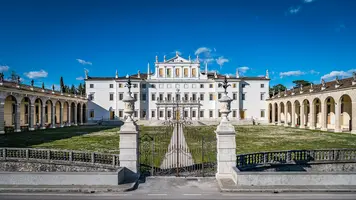 Ein prächtiges, historisches Gebäude mit mehreren Stockwerken, umgeben von einem gepflegten Garten. Die Fassade hat dekorative Elemente und eine zentrale Eingangstür. Links und rechts sind Bauten mit Bögen, die zu einem Innenhof führen. Der Himmel ist klar und blau.